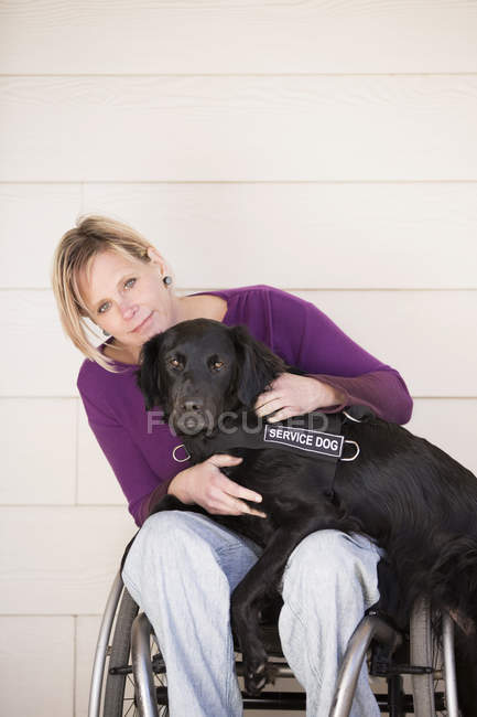 Mature woman in wheelchair hugging black service dog. — Stock Photo