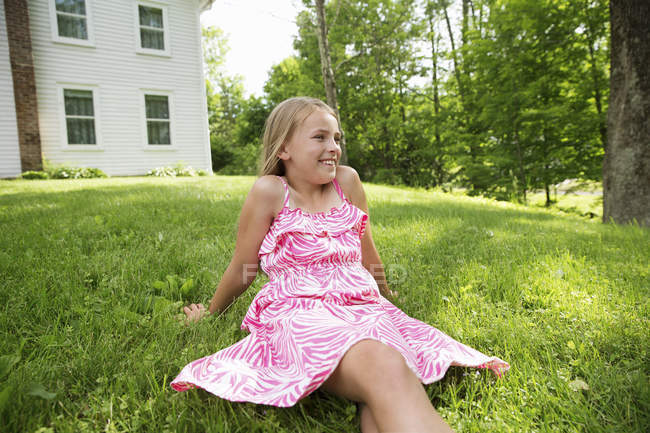 Vorpubertierendes Mädchen in pinkfarbener Dress sitzt auf Rasen im Bauerngarten. — Stockfoto