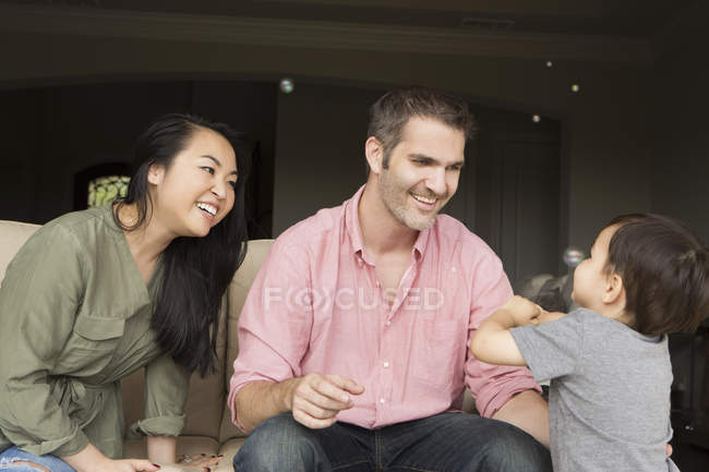 Uomo e donna sorridenti seduti fianco a fianco sul divano e che giocano con il figlio . — Foto stock
