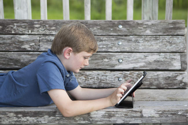Elementary age boy using digital tablet on bench outdoors. — Stock Photo