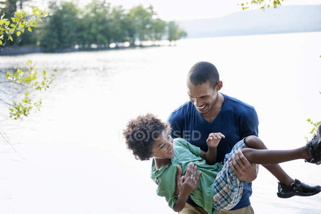 Pai brincando com filho e carregando menino em braços na margem do lago na floresta . — Fotografia de Stock