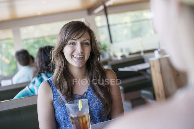Jovem mulher sentada à mesa com bebida fresca e conversando com o amigo . — Fotografia de Stock
