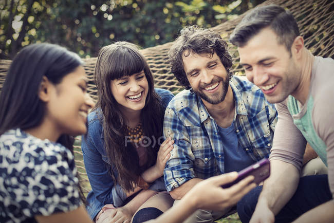 Група веселих друзів, які сидять в гамаку в саду і дивляться смартфон . — стокове фото