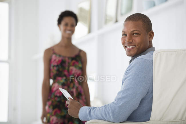 Junger Mann sitzt im Sessel und hält digitales Tablet mit Frau im Hintergrund. — Stockfoto