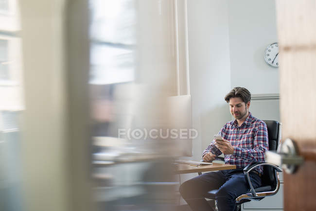 Blick durch Bürotür eines Mannes, der am Schreibtisch sitzt und sein Smartphone in der Hand hält. — Stockfoto
