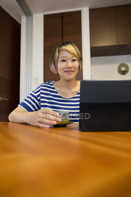 Femme japonaise assise à table avec ordinateur portable et thé vert et regardant à la caméra . — Photo de stock