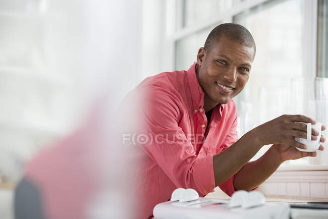 Jovem de camisa rosa segurando xícara de café e inclinando-se na soleira da janela na cozinha . — Fotografia de Stock
