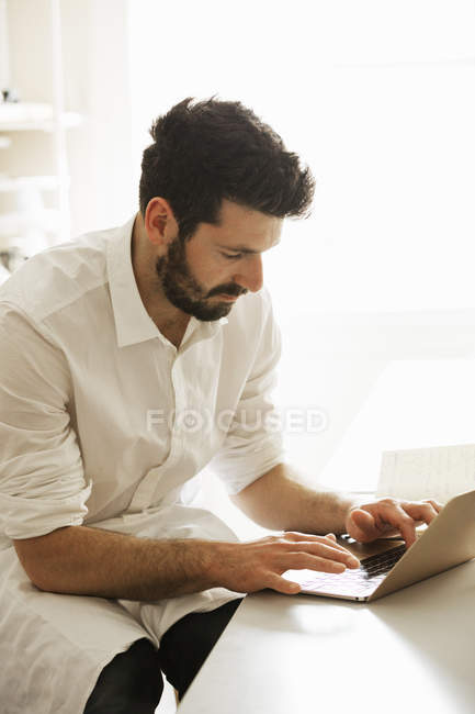 Hombre de delantal blanco sentado y escribiendo en el ordenador portátil . - foto de stock