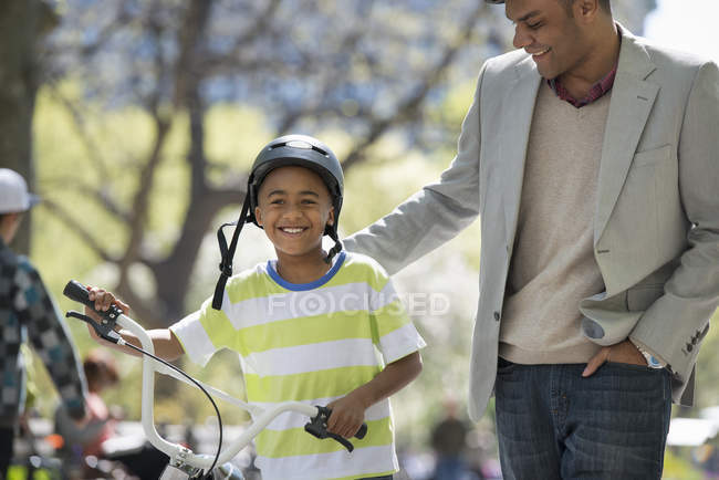Padre e figlio in casco da bicicletta a piedi con bicicletta fianco a fianco nel parco soleggiato . — Foto stock