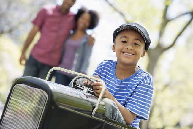 Мальчик ездит на старомодной игрушечной машине, пока родители смотрят в солнечном парке . — стоковое фото