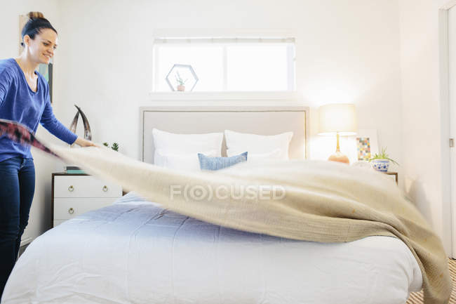 Woman spreading plain quilt across double bed in apartment bedroom. — Stock Photo
