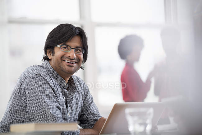Reifer Mann sitzt am Schreibtisch und benutzt Laptop im Büro, während Kollegen im Hintergrund reden. — Stockfoto