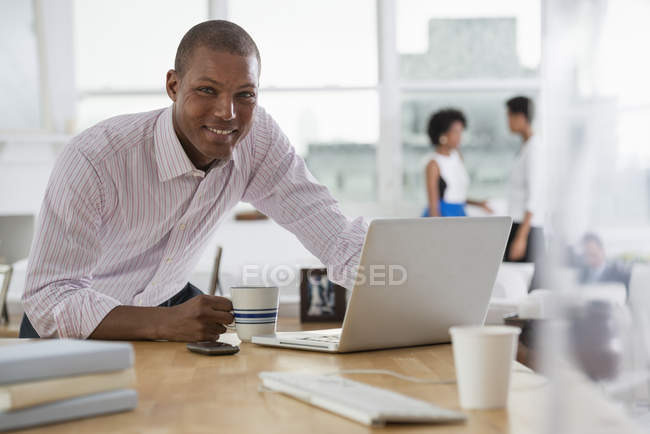 Hombre joven usando el ordenador portátil mientras se apoya en el escritorio en la oficina con compañeros de trabajo en segundo plano . - foto de stock