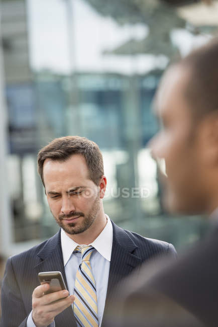 Geschäftsmann im Anzug checkt Smartphone auf der Straße mit Person im Vordergrund. — Stockfoto