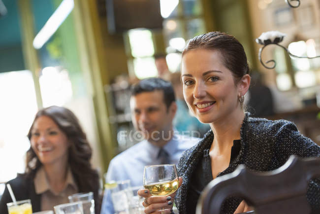 Mediados de la mujer adulta sosteniendo el vino y mirando en la cámara mientras está sentado en el bar con amigos . - foto de stock