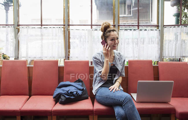 Mujer hablando por teléfono sentado en el interior en sillas con ordenador portátil . - foto de stock