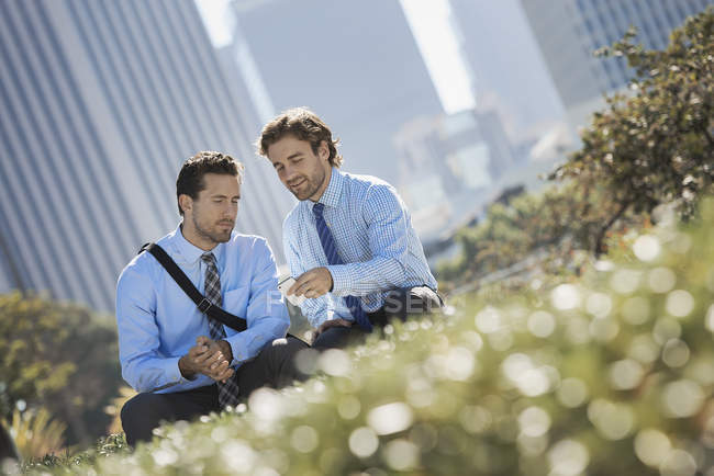 Dois homens de camisas e gravatas descansando no parque na cidade e usando telefone celular . — Fotografia de Stock
