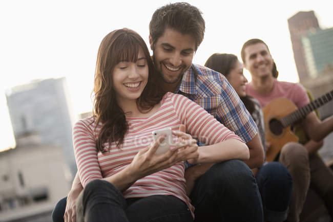 Couple using smartphone at roof party with friends and guitar. — Stock Photo