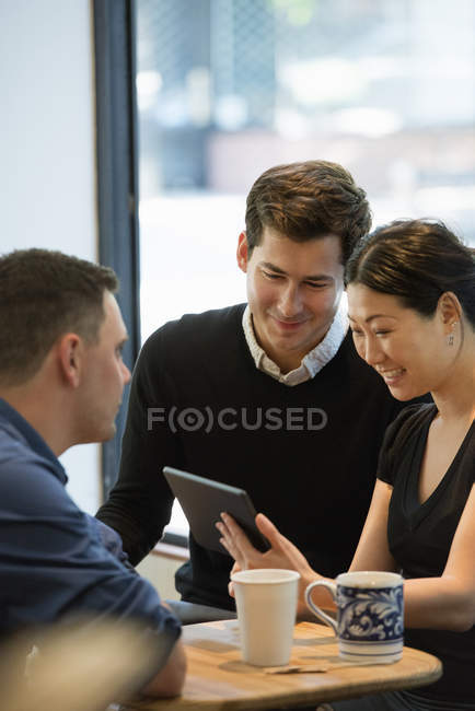 Group of friends sitting around table in coffee shop with drinks and using digital tablet. — Stock Photo