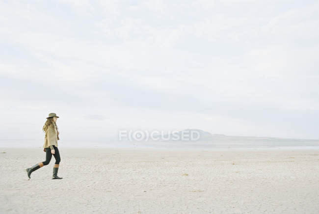 Mulher com cabelos longos e encaracolados andando na praia de areia usando chapéu e botas de couro . — Fotografia de Stock