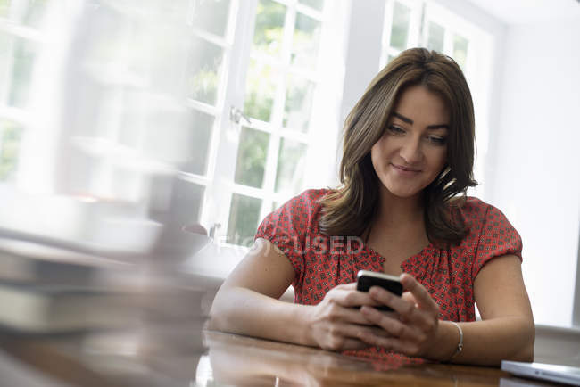Жінка сидить за столом в приміщенні і перевіряє смартфон . — стокове фото