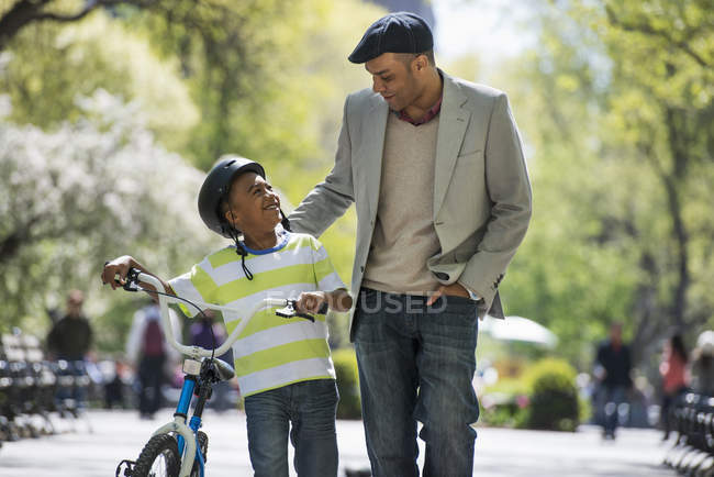 Padre e hijo en casco de bicicleta caminando con bicicleta lado a lado en el soleado parque . - foto de stock
