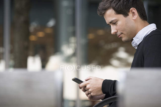 Vista lateral do jovem sentado no banco e usando smartphone . — Fotografia de Stock