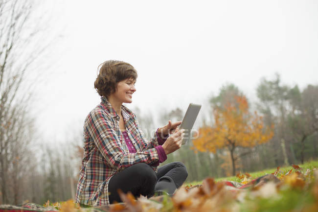 Mitte erwachsene Frau sitzt mit digitalem Tablet auf dem Boden im herbstlichen Wald. — Stockfoto