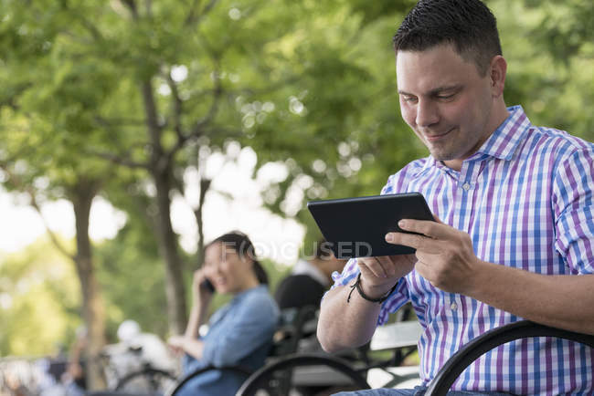 Man using digital tablet on bench in city park with woman talking on phone in background. — Stock Photo