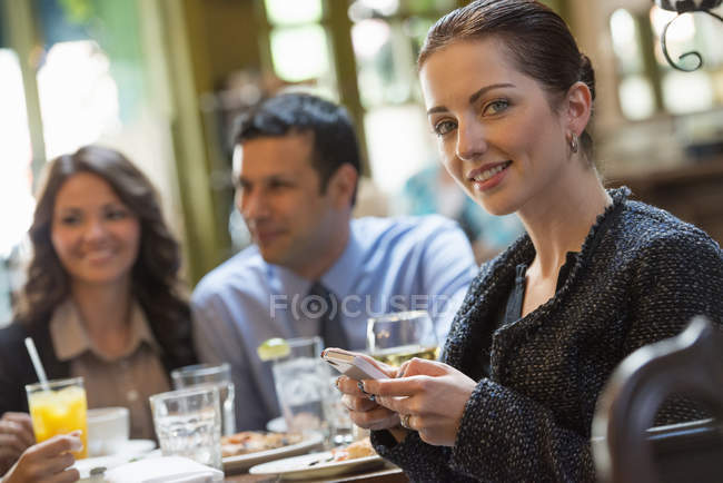 Mujer adulta media sosteniendo el teléfono inteligente y mirando en la cámara mientras está sentado en el bar con amigos . - foto de stock