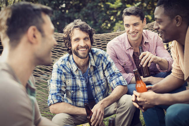 Group of cheerful male friends lounging in hammock in garden and drinking beer. — Stock Photo