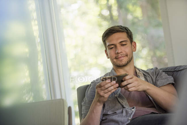 Мужчина сидит на диване и проверяет смартфон в помещении . — стоковое фото
