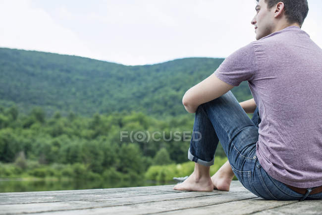 Giovane seduto su un molo di legno vicino al lago in estate — Foto stock