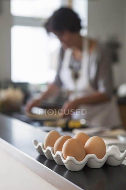 Tray of chicken eggs on kitchen table with woman cooking in background. — Stock Photo