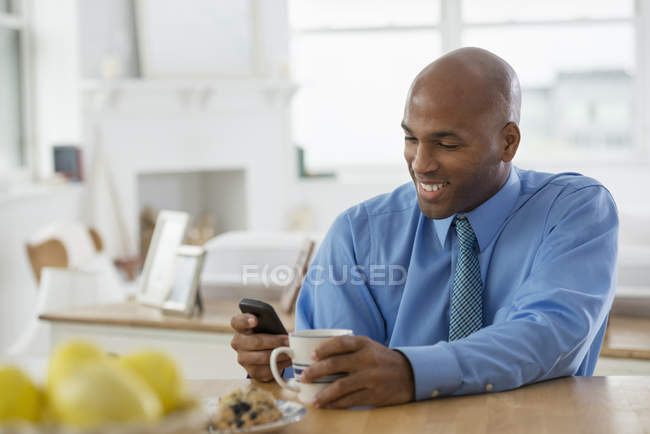 Uomo in camicia blu seduto al tavolo della colazione con caffè e utilizzando smartphone
. — Foto stock