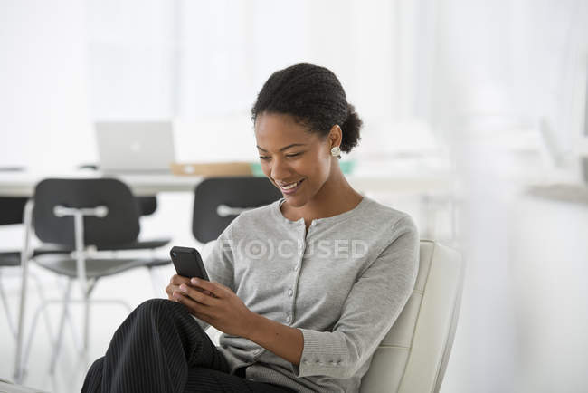 Femme d'affaires joyeuse utilisant un smartphone dans une chaise confortable au bureau
. — Photo de stock