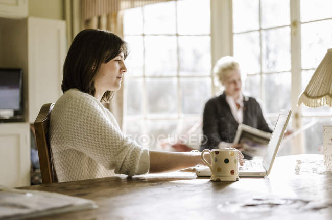 Frau arbeitet mit Laptop am Küchentisch mit Seniorin im Hintergrund. — Stockfoto