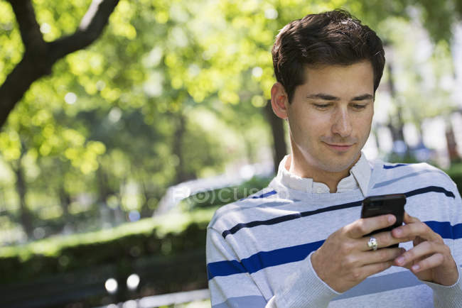 Jeune homme vérifiant smartphone tout en étant assis dans le parc de la ville . — Photo de stock