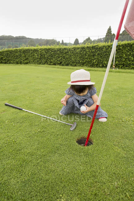 Menina da criança sentada perto do buraco do campo de golfe e segurando bola . — Fotografia de Stock