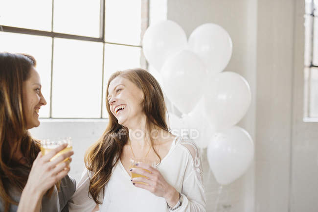 Two women standing side by side and holding drinks in room decorated with white balloons. — Stock Photo