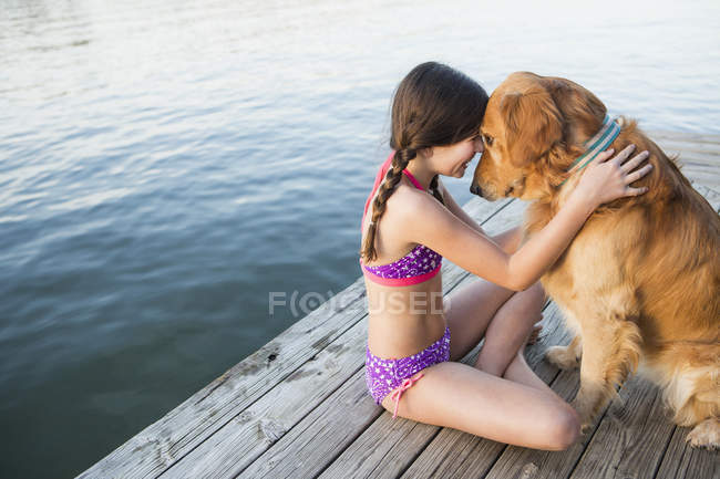 Pre-adolescent girl in swimwear with golden retriever dog sitting on jetty. — Stock Photo