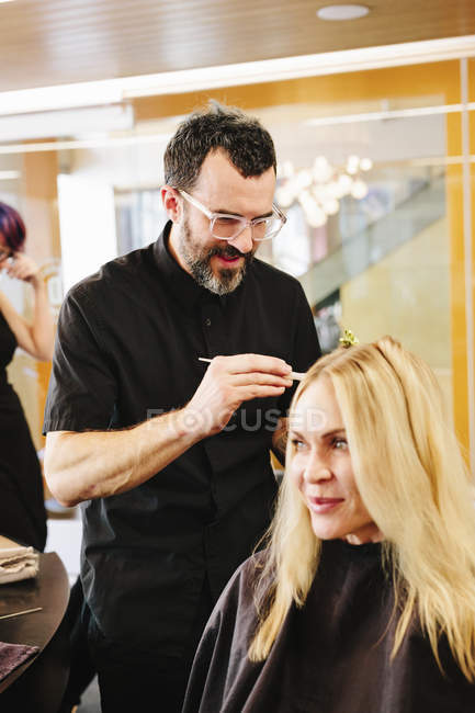 Coiffeur masculin mature travaillant sur les cheveux du client et appliquant une teinture capillaire dans le salon . — Photo de stock