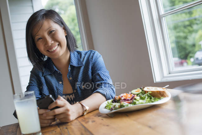 Азиатка держит смартфон, сидя за столом с кофе и салатом . — стоковое фото