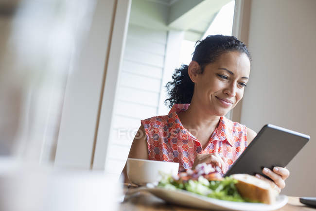 Woman smiling while using digital tablet at cafe table with coffee and sandwich. — Stock Photo