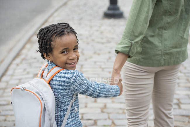 Niño con bolsa de libro caminando de la mano con la mujer y mirando hacia atrás en la cámara . - foto de stock