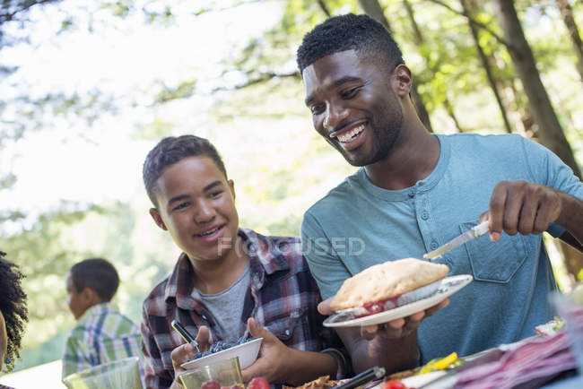 Young man and boy cutting fruit pie at picnic table. — Stock Photo