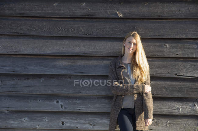 Young woman leaning against wooden barn and looking in camera in winter. — Stock Photo