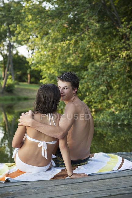 Young man looking over shoulder while sitting with young woman on wooden jetty by water. — Stock Photo