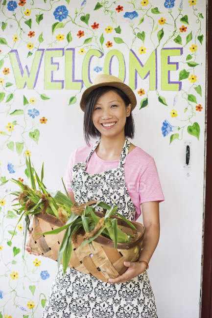 Young Asian woman standing by Welcome sign with basket of freshly picked green farm vegetables. — Stock Photo