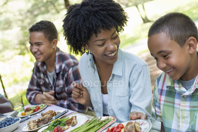 Mulher e meninos sentados na mesa de piquenique com comida e rindo . — Fotografia de Stock